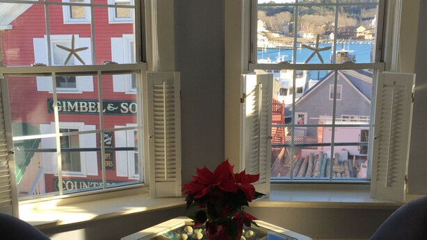 Una habitación con dos ventanas, una mesa con un florero y vista a un edificio rojo al otro lado de la calle.