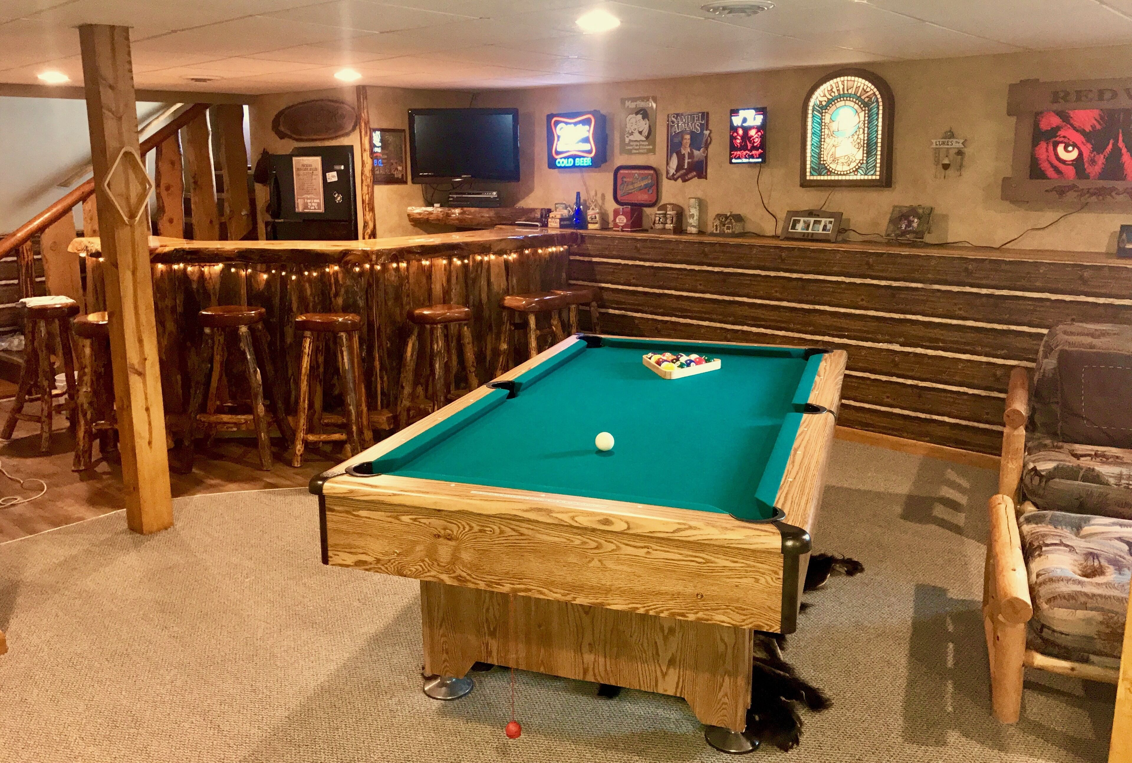 Une table de billard dans une pièce comprenant un bar, des tabourets en bois et un escalier.