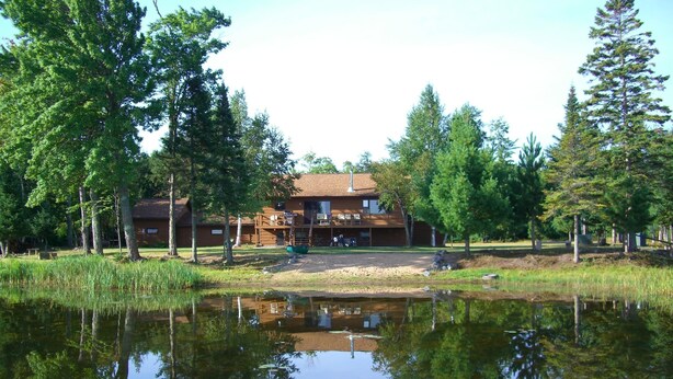 A two-story lodge with a large deck and outdoor seating area, surrounded by trees and a pond.
