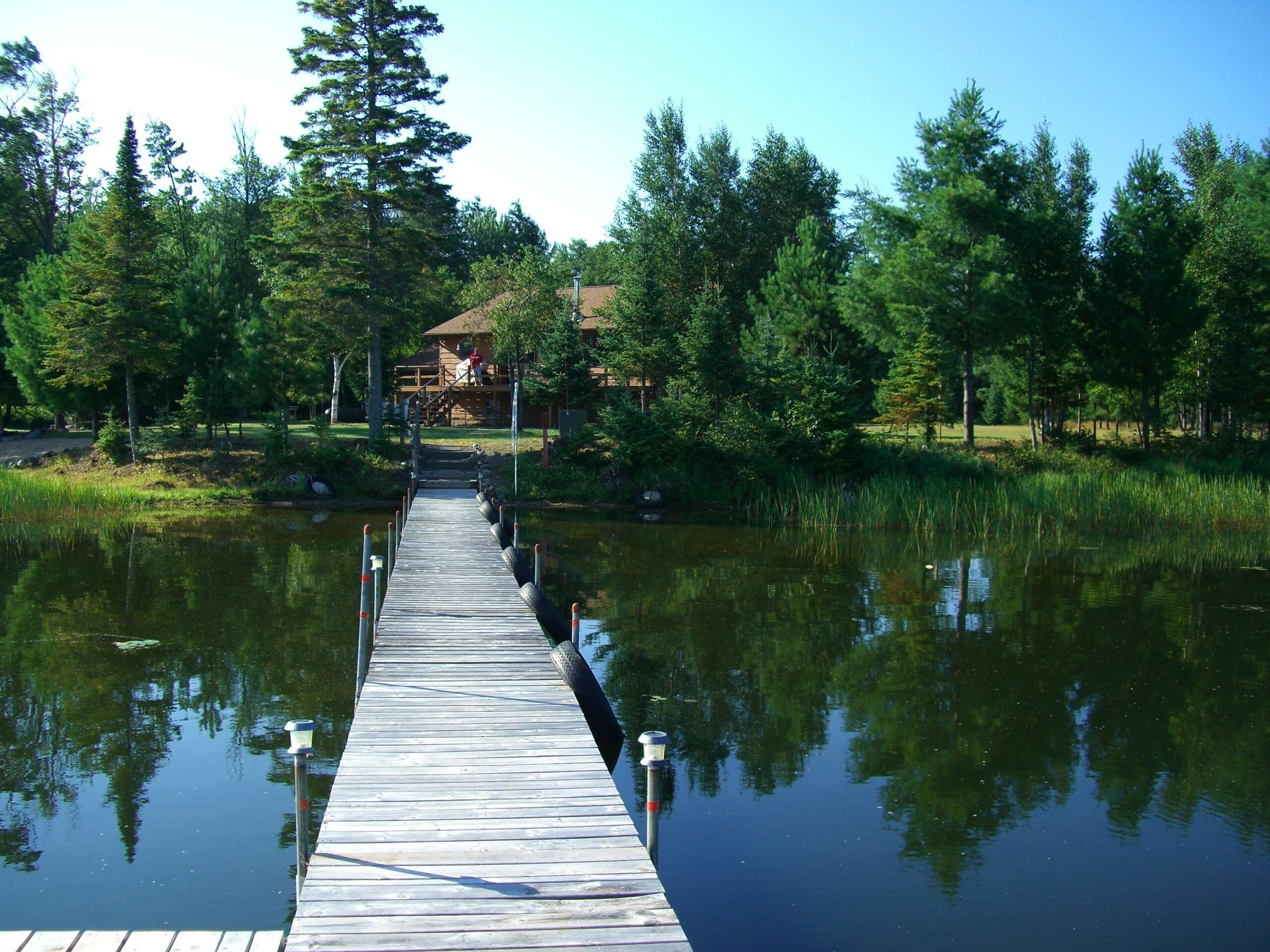 A wooden pier extending into a calm lake, surrounded by dense trees and a lodge in the background.