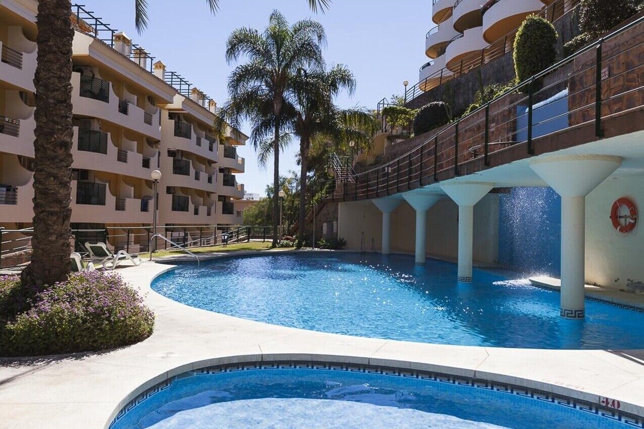 En swimmingpool omgivet af et hotel med mange etager, altaner, palmer og en klar himmel.