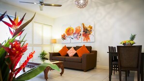 A living room with a brown sofa, orange cushions, a dining table with chairs, a ceiling fan, and a wall-mounted abstract painting. - 5313 Coral Suite The Beach Club (Palm Cove)