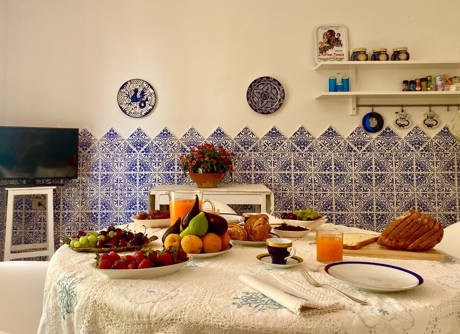 A breakfast table with fruits, bread, and drinks, set against a blue and white tiled backsplash.