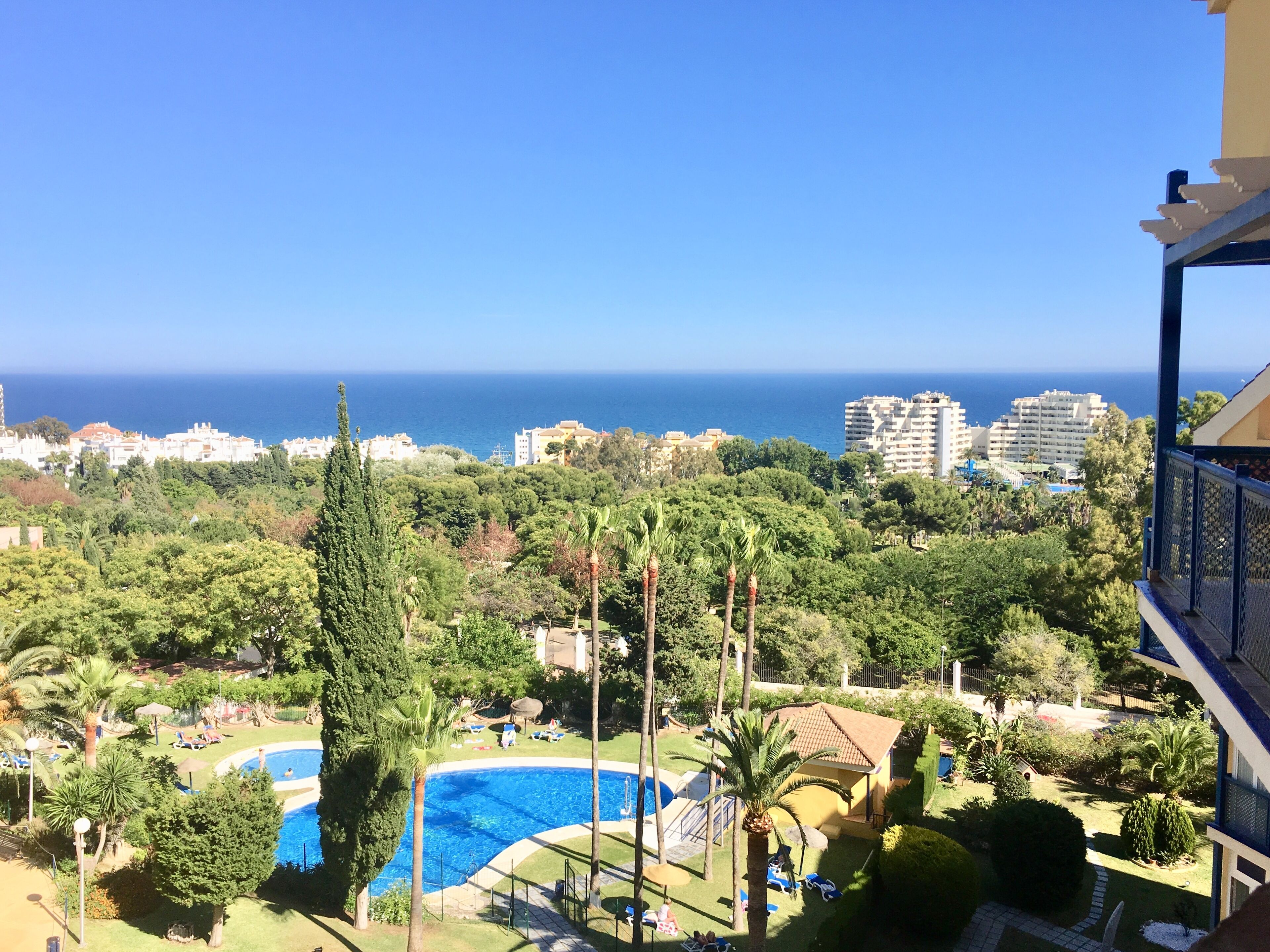 A view of a resort with a swimming pool, lush greenery, and a clear blue sea in the background.