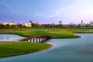 Ein Golfplatz mit einer Holzbrücke über einem Wasserspiegel, umgeben von Grünflächen und Gebäuden im Hintergrund.