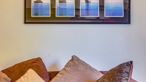 A room with a brown leather sofa and a framed picture of a sunset over water.