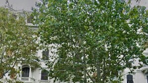 A multi-story building with a classical architectural style, featuring arched windows and balconies. There is a tree in front of the building, and a white van parked on the street.