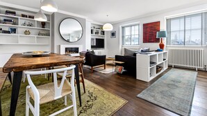 A modern living room with a wooden dining table, white chairs, a black sofa, and a white bookshelf.