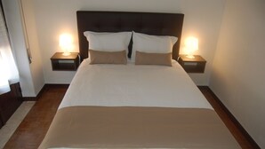 Ein ordentlich bezogenes Bett mit weißer und beiger Bettwäsche, zwei Nachttische mit Lampen und ein Holzboden.