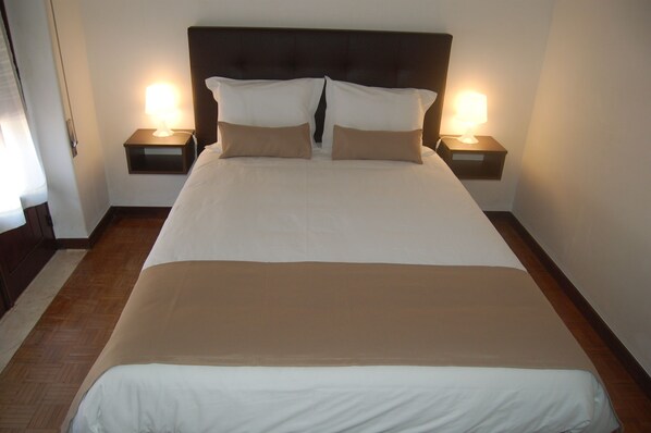 Una cama bien hecha con ropa de cama blanca y beige, dos mesitas de noche con lámparas y suelo de madera.