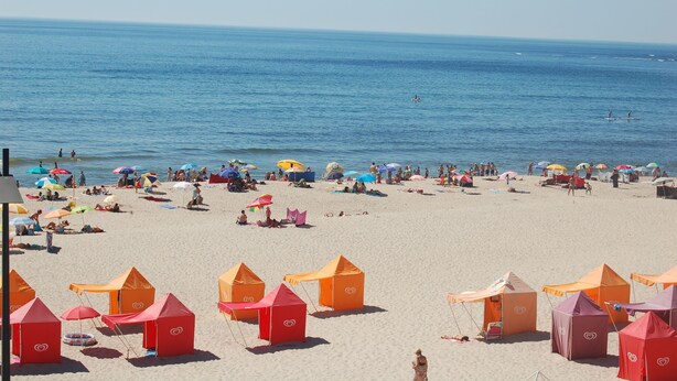 Ein Strand mit Zelten und Sonnenschirmen, entspannte Menschen und das Meer im Hintergrund.