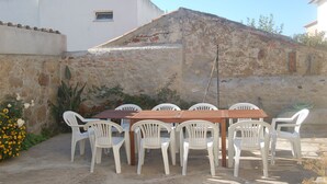 Eine Terrasse mit Holztisch und weißen Plastikstühlen, vor einer Steinmauer und einem Gebäude mit weißem Putz und roten Ziegeln.
