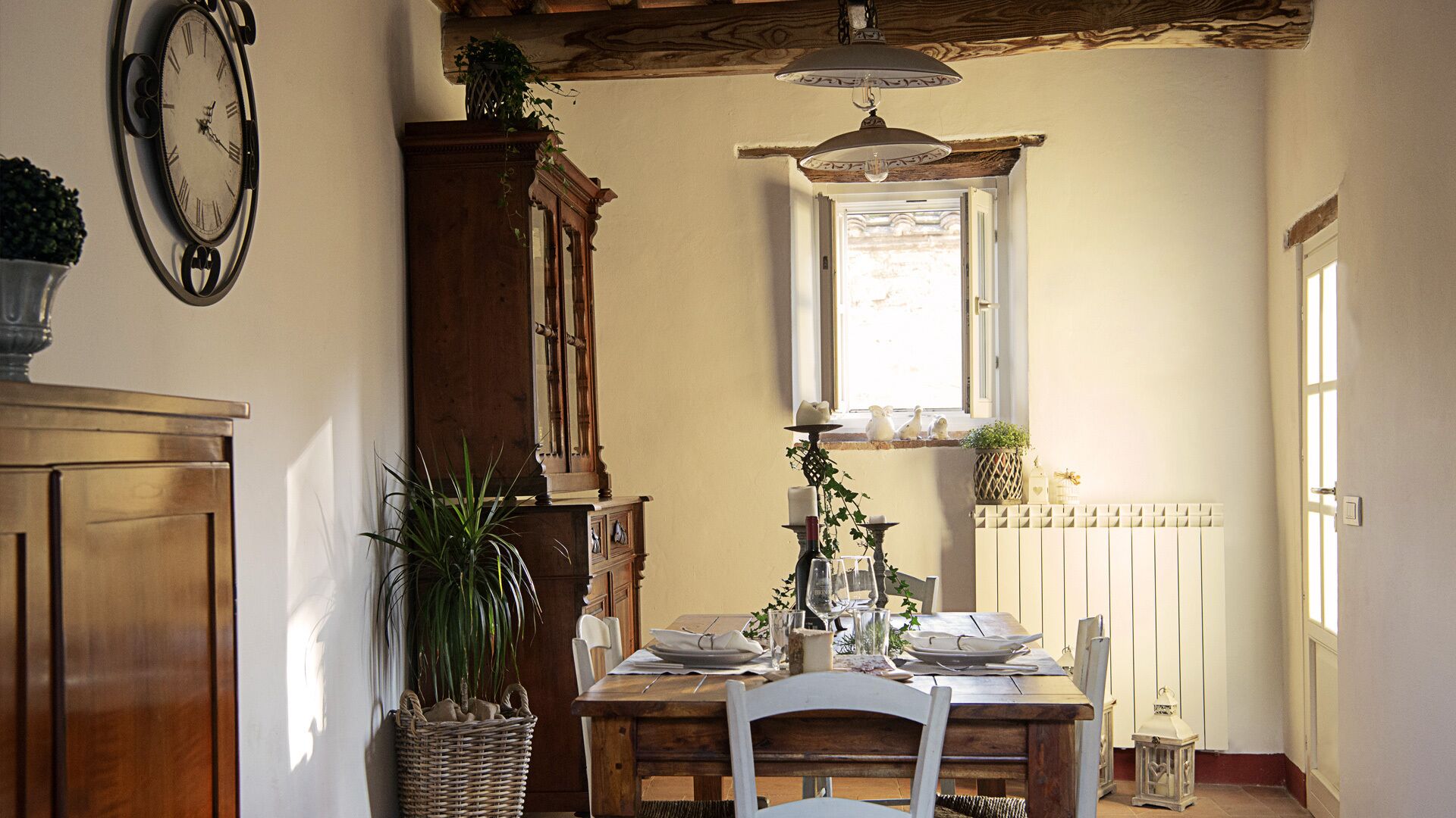 Un comedor con una mesa de madera preparada para dos personas, un reloj grande en la pared y una ventana con vistas al exterior.