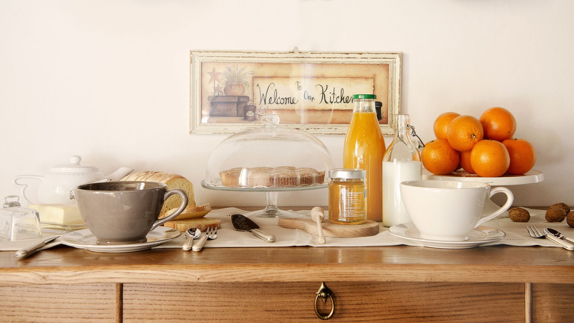 Frühstückstisch mit einer Kaffeetasse, Teekanne, Brot, Orangen und einem Schild mit der Aufschrift 'Welcome to Our Kitchen'.