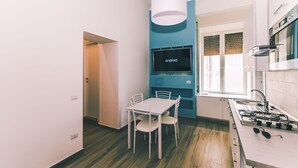 A modern kitchen with a built-in stove, a dining table with chairs, a TV mounted on the wall, and a window with blinds.