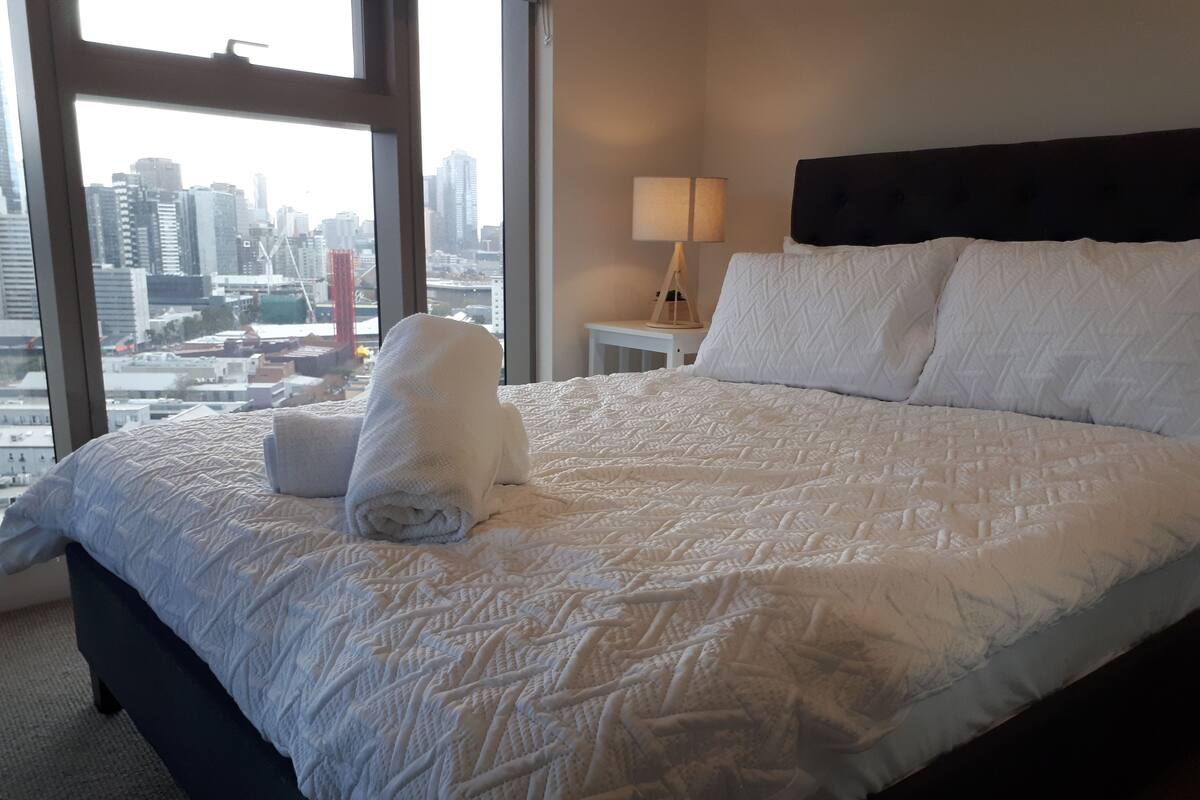 A neatly made bed with a white quilt, a rolled white towel, and a nightstand with a lamp.