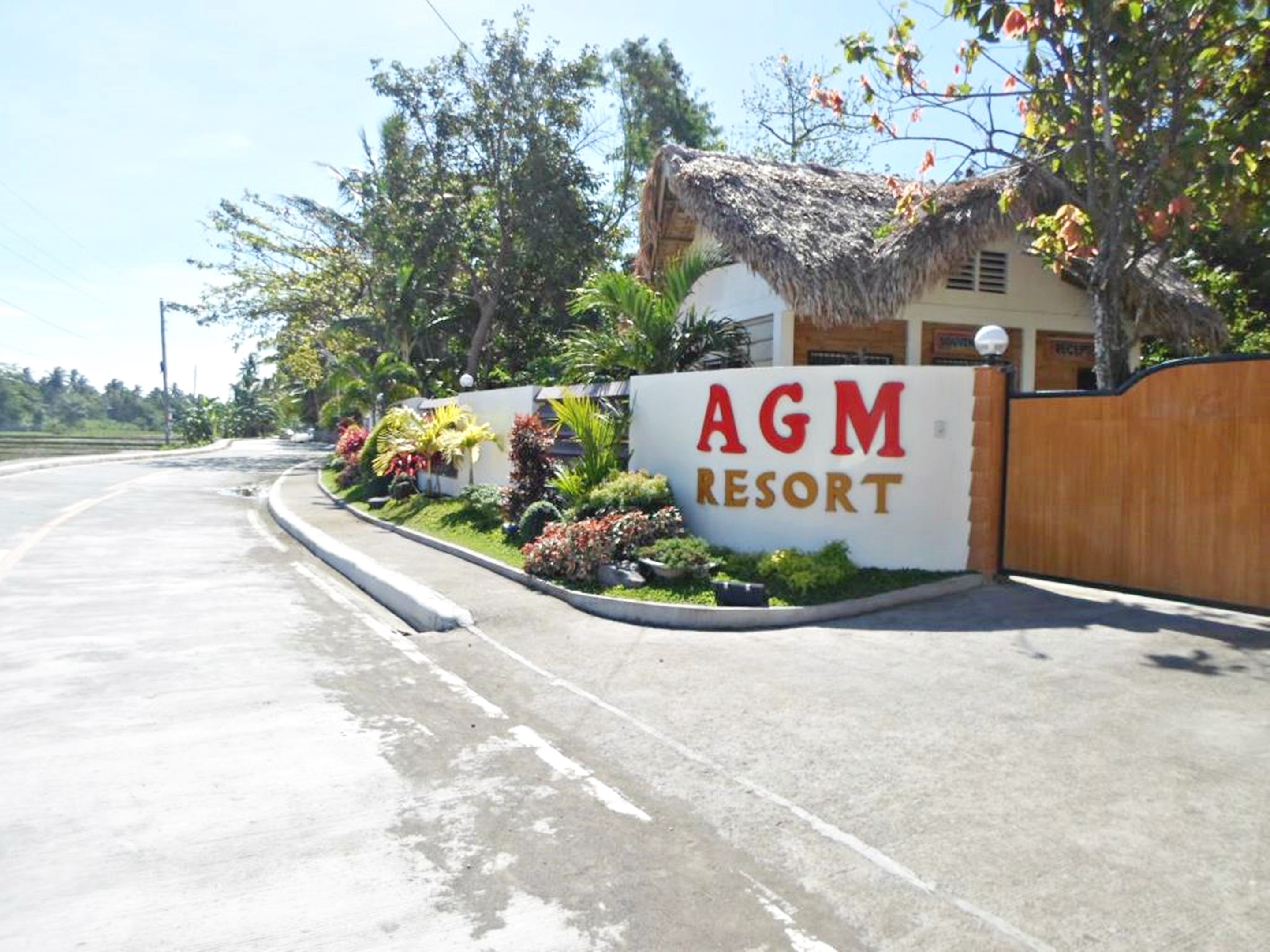 Señal de AGM Resort con un edificio de techo de paja y jardín ajardinado.