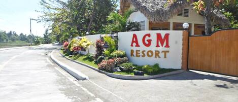 Señal de AGM Resort con un edificio de techo de paja y jardín ajardinado.