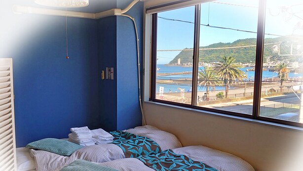 Une chambre avec un lit adossé à un mur bleu, une fenêtre donnant sur une zone côtière et un plafond blanc.