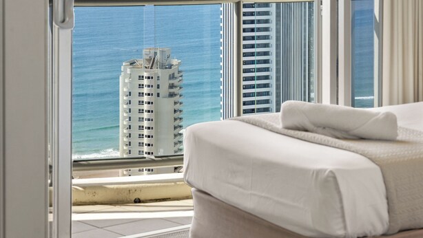 Habitación de hotel con un amplio ventanal corredera que da a un balcón con vistas al océano y edificios altos.