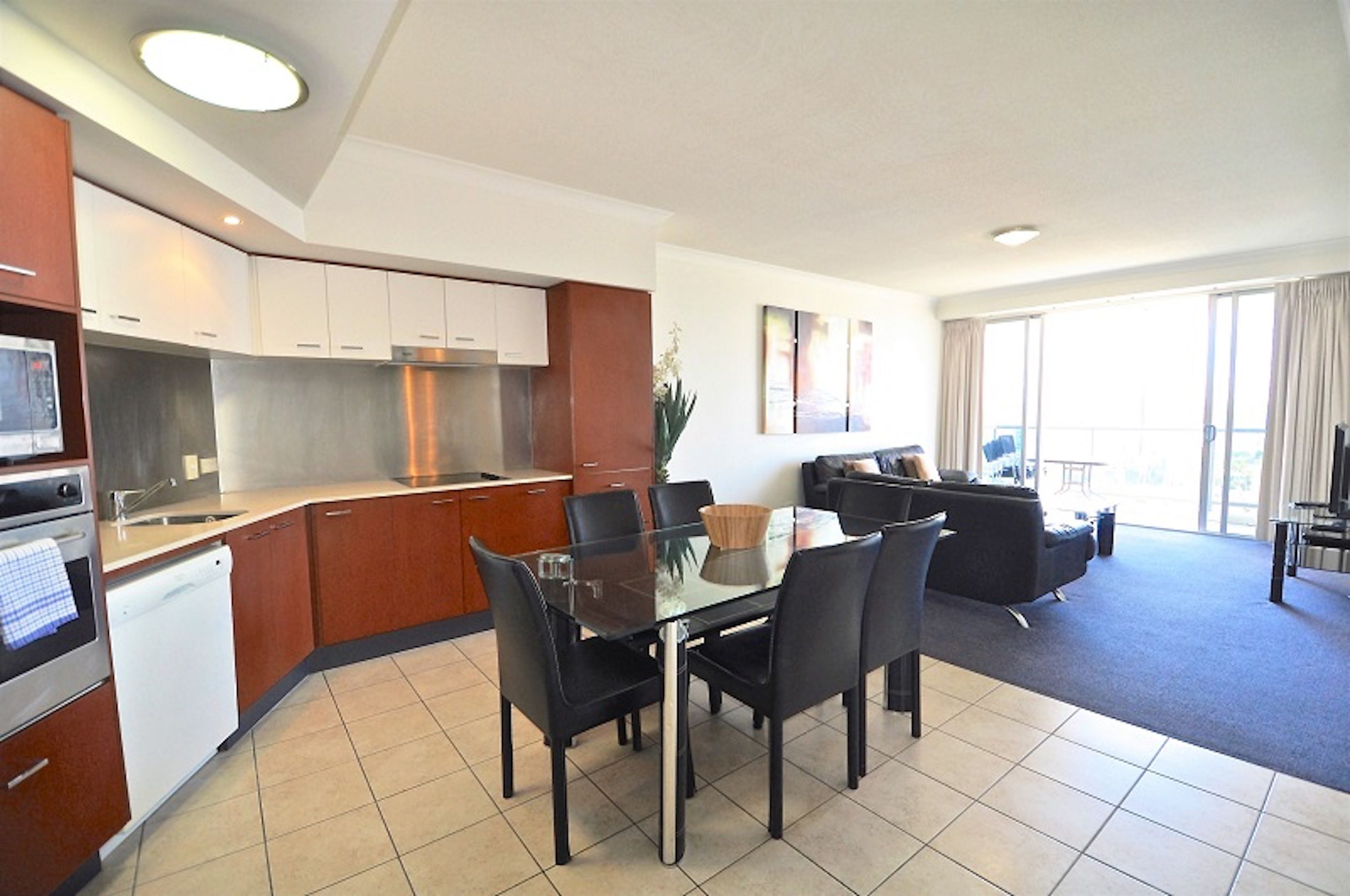 Una cocina moderna con electrodomésticos de acero inoxidable, un comedor con una mesa de vidrio y una sala de estar con sofá y televisor.