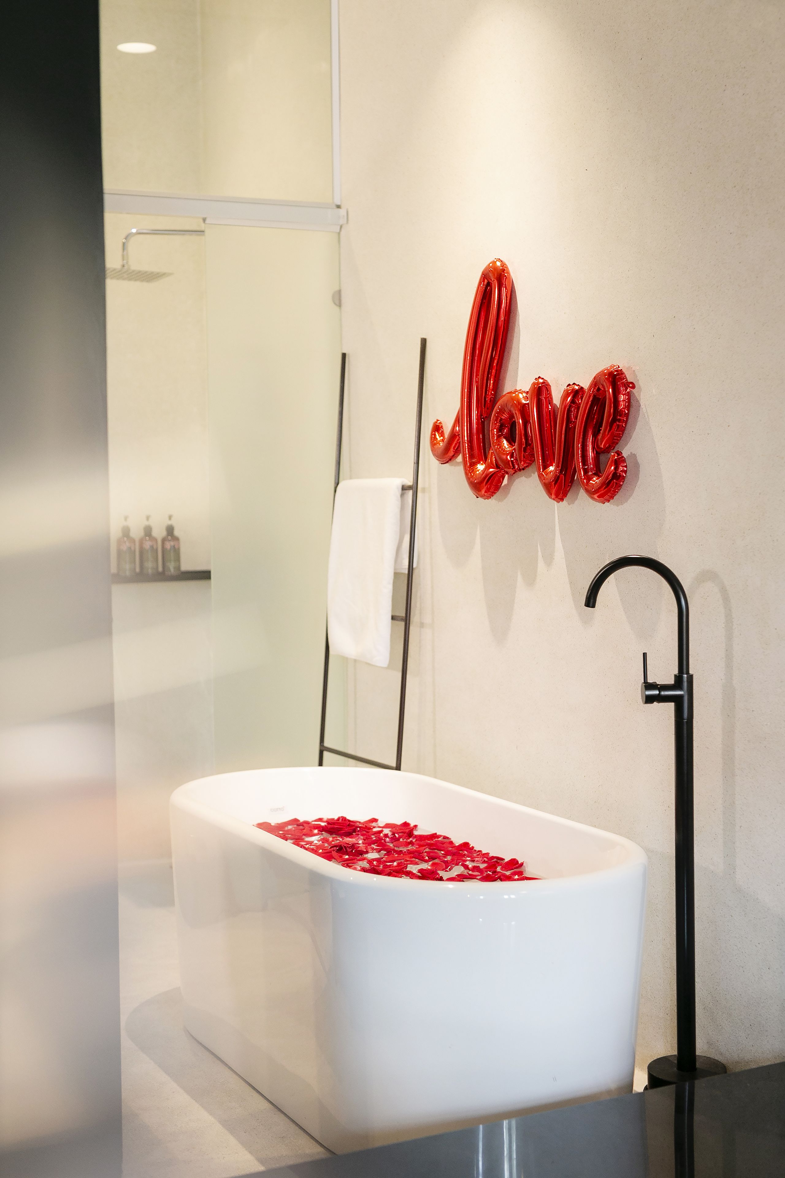 A modern bathroom with a white bathtub filled with rose petals, a black faucet, and a red "Love" sign on the wall.