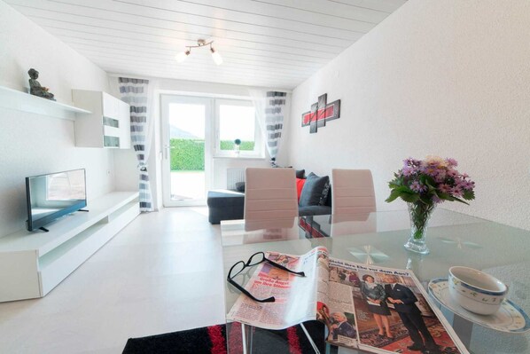 A modern living room with a glass table, a sofa, a TV, and a window with curtains.