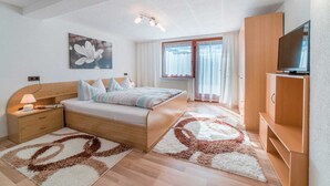 A bedroom with a bed, bedside table, lamp, TV, wooden wardrobe, and a large window with curtains.