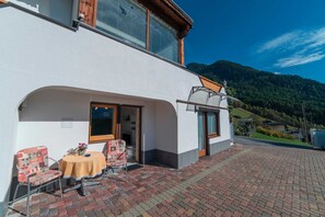 A two-story building with a patio area featuring a table and chairs, a covered outdoor seating area, and a mountain backdrop.