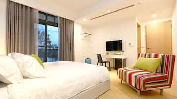 A hotel room with a large bed, a desk with a TV, a colorful sofa, and a view of trees through the window.