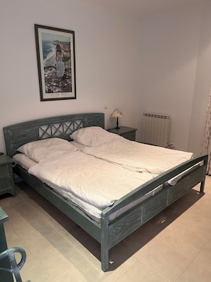Een bed met houten frame, een nachtkastje met lamp, een ingelijst beeld aan de muur en een radiator.