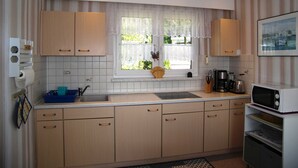 Een keuken met houten kastjes, een raam met kantgordijnen, een spoelbak en een magnetron.