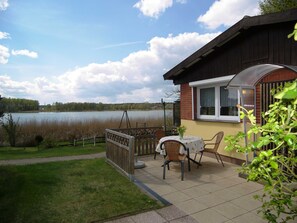 Een terras met tafel en stoelen met uitzicht op een meer, een houten hek en een gebouw met een raam.