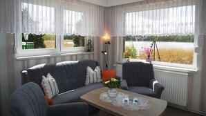 Een gezellige kamer met een grijze bank, twee fauteuils, een houten bijzettafeltje met een kant kleedje, en uitzicht op een meer door grote ramen met dunne gordijnen.