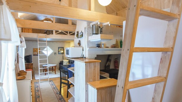 Interior rústico de una cabaña con cama en un entrepiso, comedor y cocina con estantes de madera.
