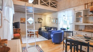 Una sala de estar acogedora con techo de madera, un sofá, un sillón azul, una mesa de comedor con sillas y una escalera que sube a un nivel superior.