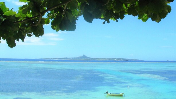 A coastal landscape with clear turquoise waters, a small boat, and a distant mountain. - Fukugi Terrace (Motobu)