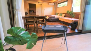A room with a bed, a dining table with chairs, a grill, and a large plant. - Fukugi Terrace (Motobu)