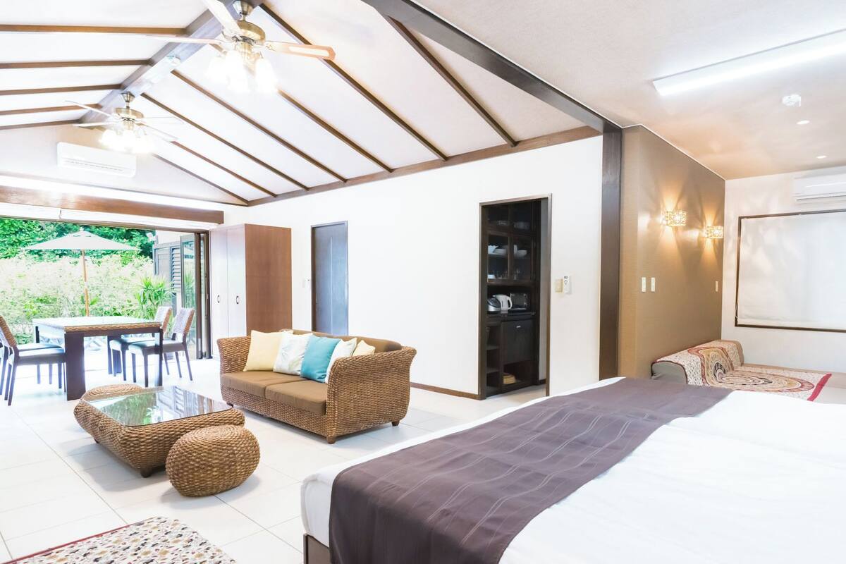 A bedroom with a bed, a sofa, a dining area with chairs, and a view of the outdoors.