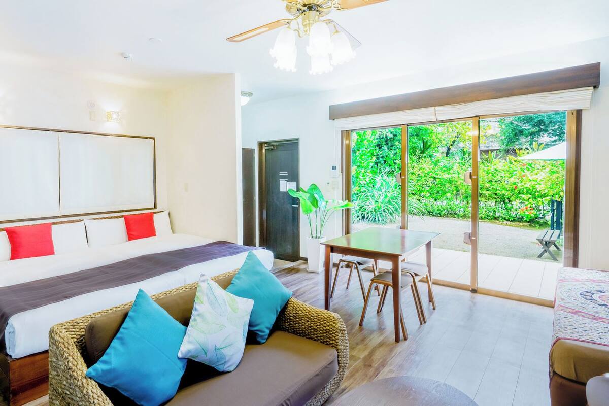 A bedroom with a bed, a sofa, a dining table, and a view of greenery outside.