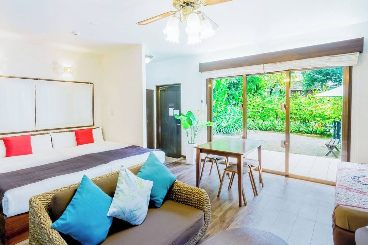 A bedroom with a bed, a sofa, a dining table, and a view of greenery outside.