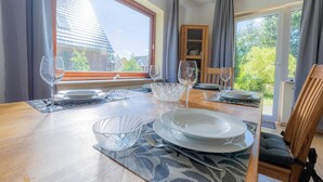 Zona pranzo con tavolo di legno apparecchiata per quattro persone, ampie finestre e vista su una casa e del verde all'esterno.
