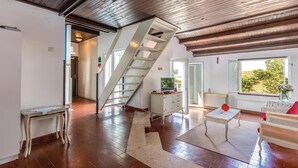 Ein geräumiges Zimmer mit Holzboden, einer Treppe, einem Fernseher und einem Fenster mit Blick ins Grüne.