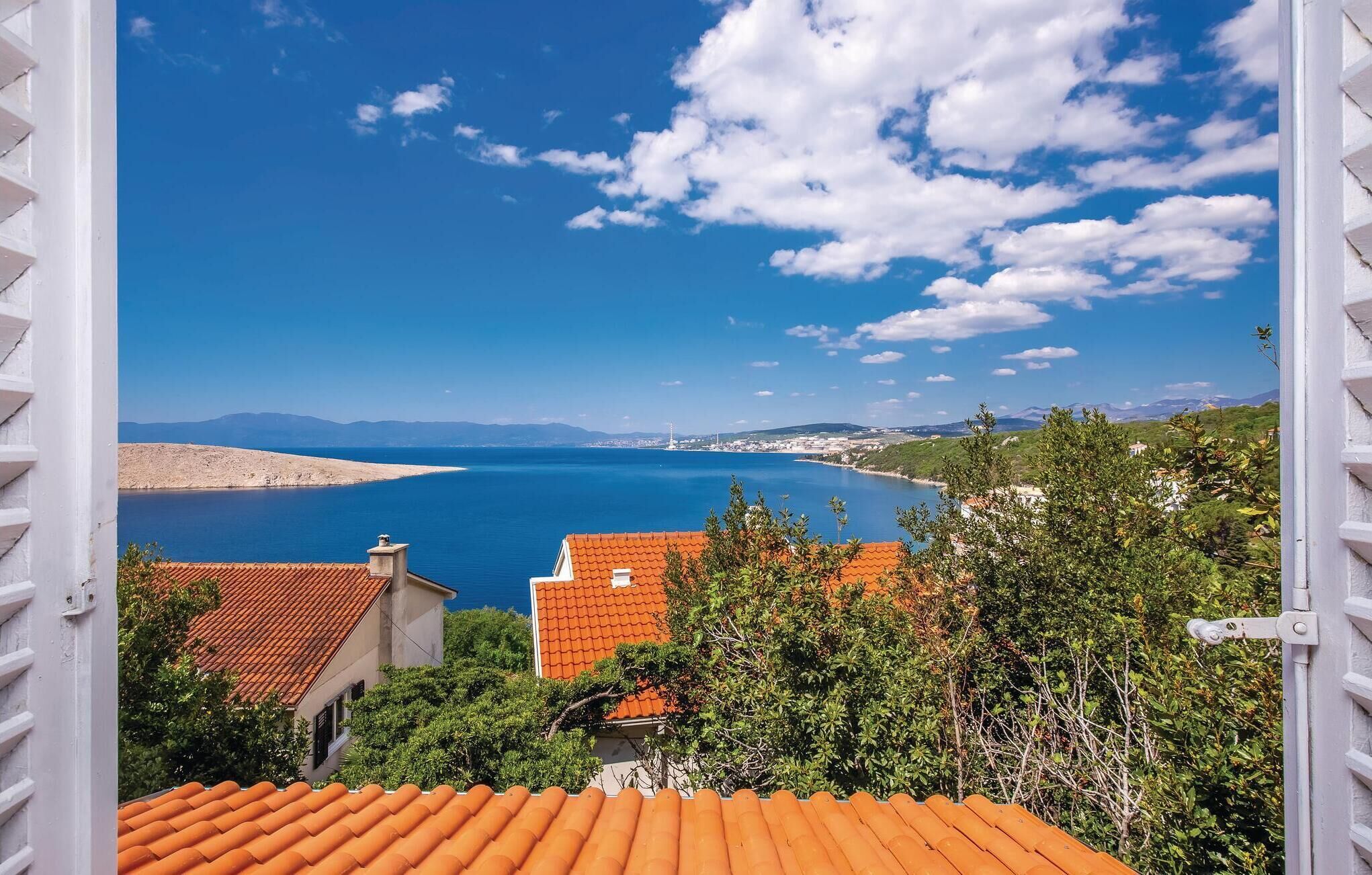 A coastal view from a balcony with red-tiled roofs, overlooking a calm sea and distant mountains.