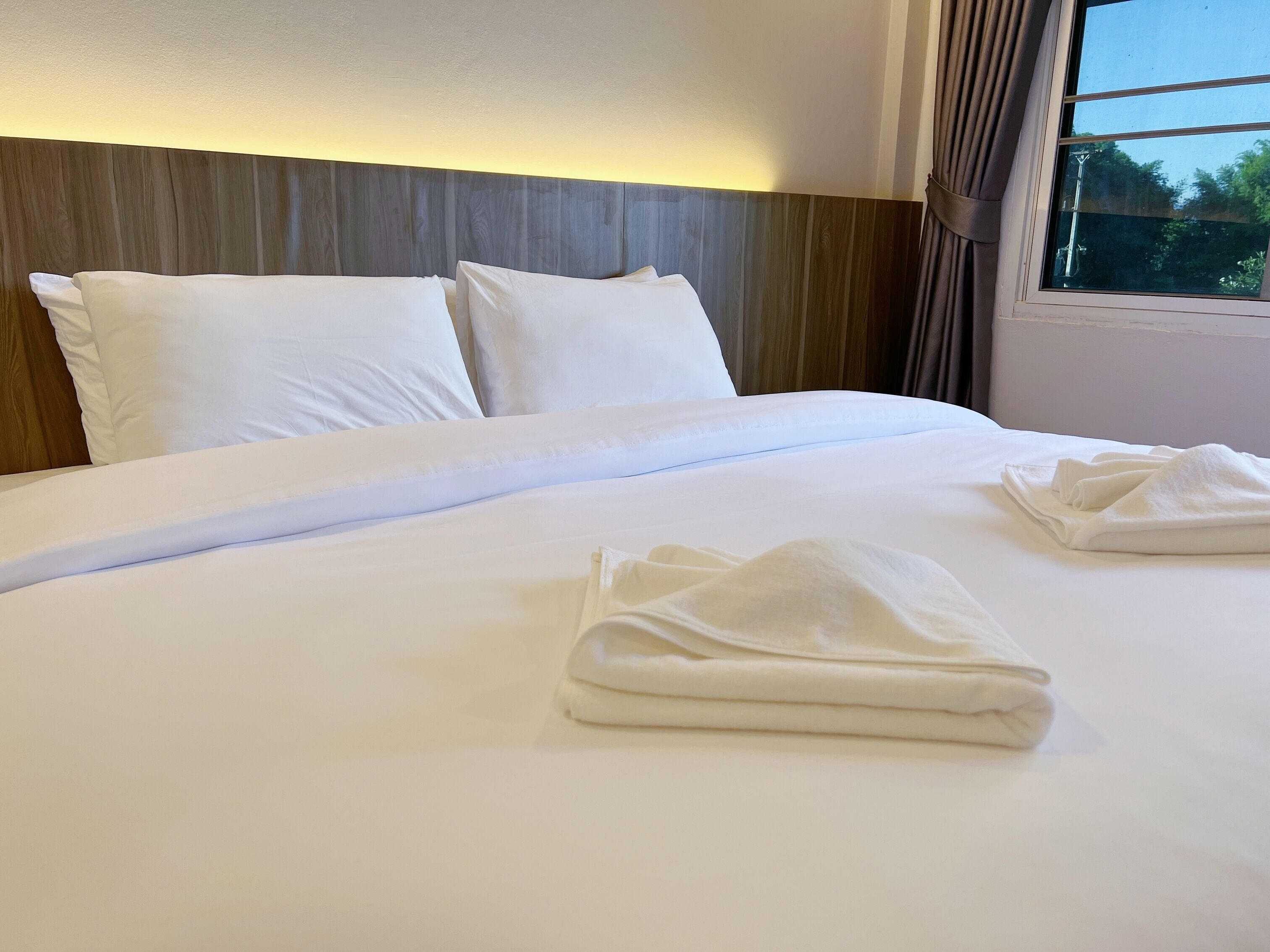 A neatly made bed with white linens and two folded towels, a wooden headboard, and a window with curtains.