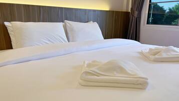 A neatly made bed with white linens and two folded towels, a wooden headboard, and a window with curtains.