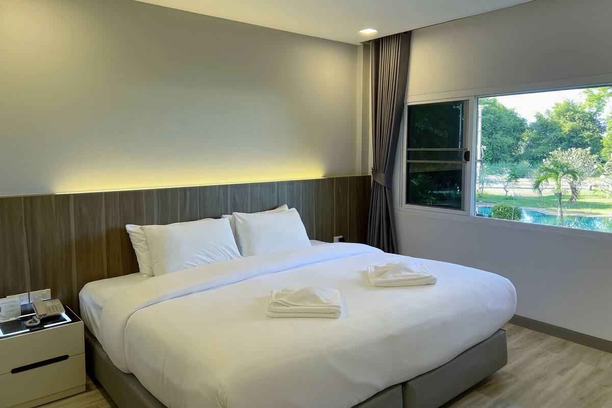A modern hotel room with a large bed, a bedside table with a phone, and a window with a view of a swimming pool and greenery.