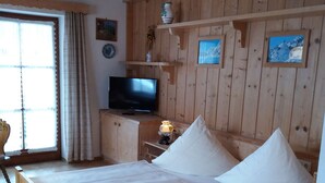A wooden-paneled room with a bed, a television, and a window with curtains. - Vacation apartment Schönau 4 pers., 1 bedroom, 1 bed-sitting room, balcony, 48 sqm (Inzell)