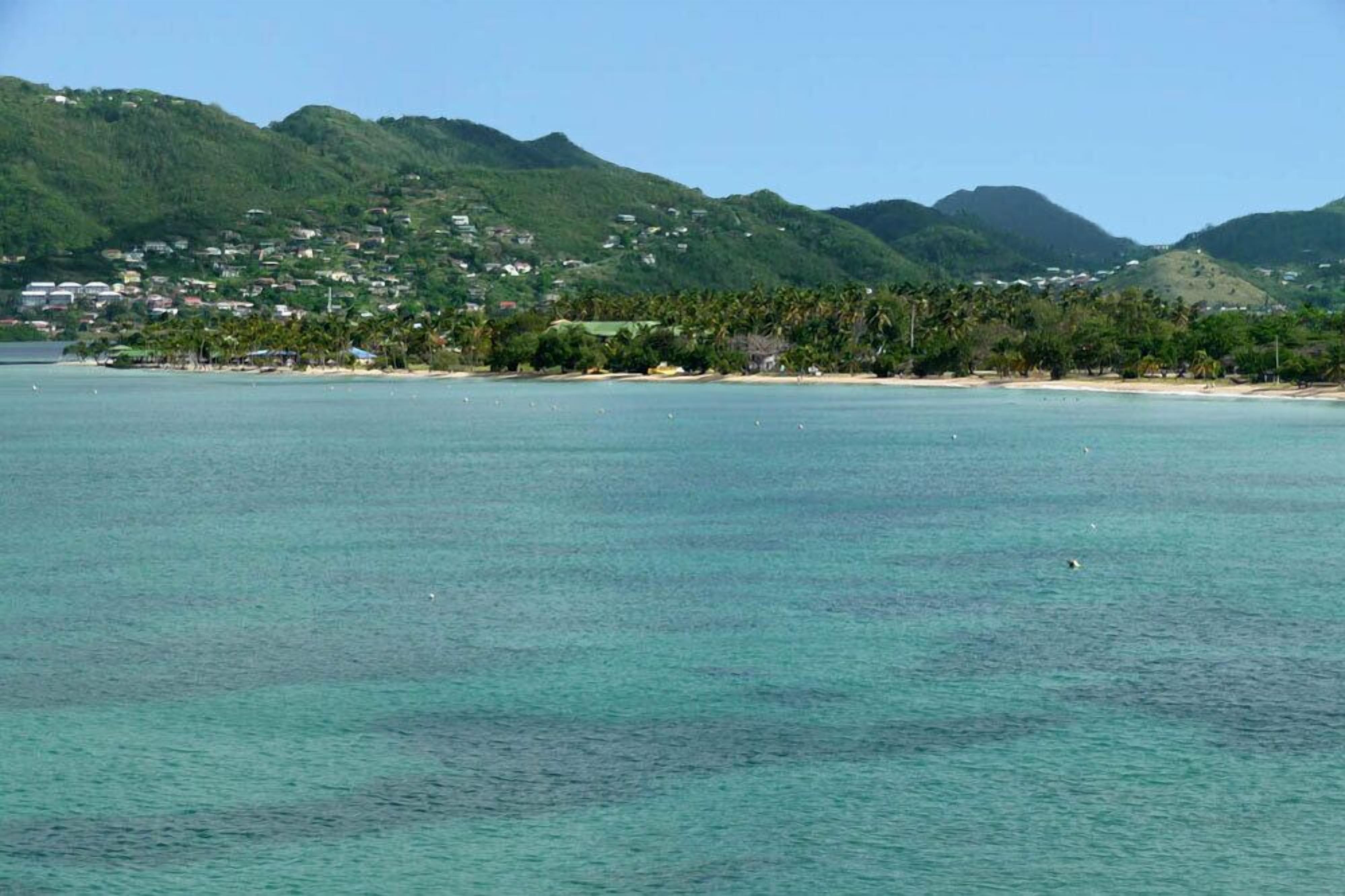 A coastal landscape with a beach, clear turquoise water, and a backdrop of green hills with scattered buildings.