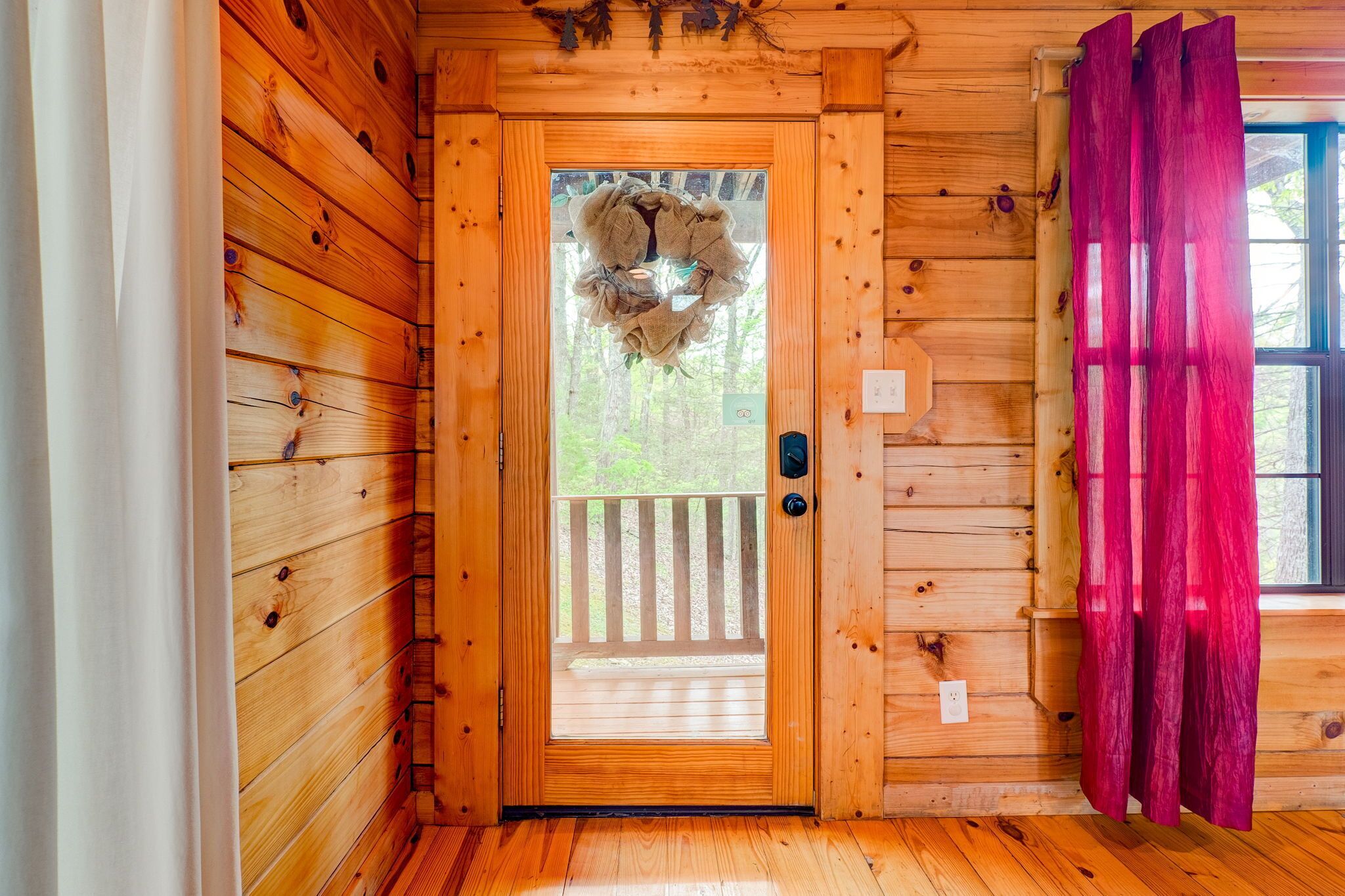 Eingang einer Holzjagdhuette mit Glastür, dahinter ein Holzdeck und ein roter Vorhang auf der rechten Seite.